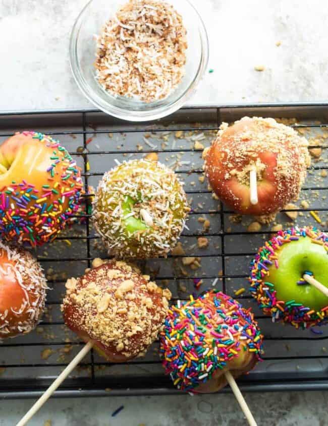 A variety of candy apples on a cooling rack, coated with colorful sprinkles, crushed nuts, and coconut flakes showcase an irresistible treat from your favorite snack recipes. Bowls with extra toppings are scattered around on a light textured surface.