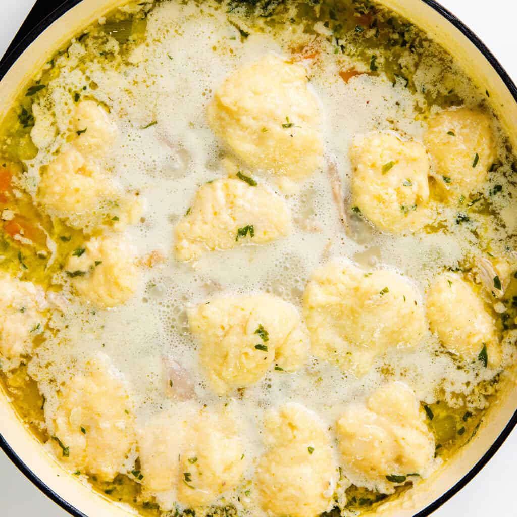 A pot filled with homemade chicken dumpling soup. The golden dumplings float in a creamy broth with visible herbs and pieces of chicken. The dish appears hearty and comforting, exuding a rich, savory aroma that promises warmth with every spoonful.