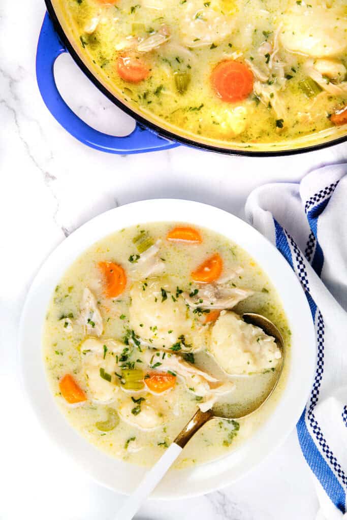 A bowl of creamy chicken dumpling soup with carrots, celery, and herbs sits on a white marble surface. Beside it is a blue pot brimming with more of this heartwarming chicken recipe. A white and blue dish towel adds a cozy touch to the scene on the right.