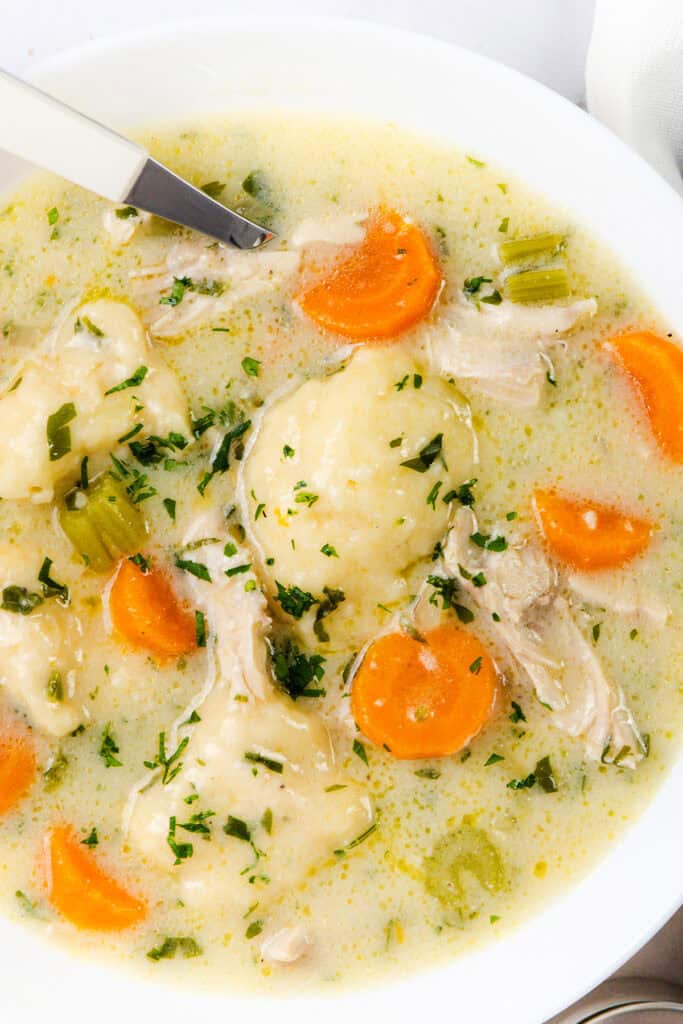 A comforting bowl of creamy chicken and dumpling soup features sliced carrots, celery, shredded chicken, and fluffy dumplings. Garnished with chopped parsley and served with a white-handled spoon.