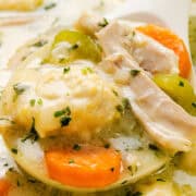 A close-up of a spoonful of hearty chicken and dumpling soup reveals tender chicken chunks, fluffy dumplings, sliced carrots, and peas in a creamy broth, all garnished with fresh herbs.