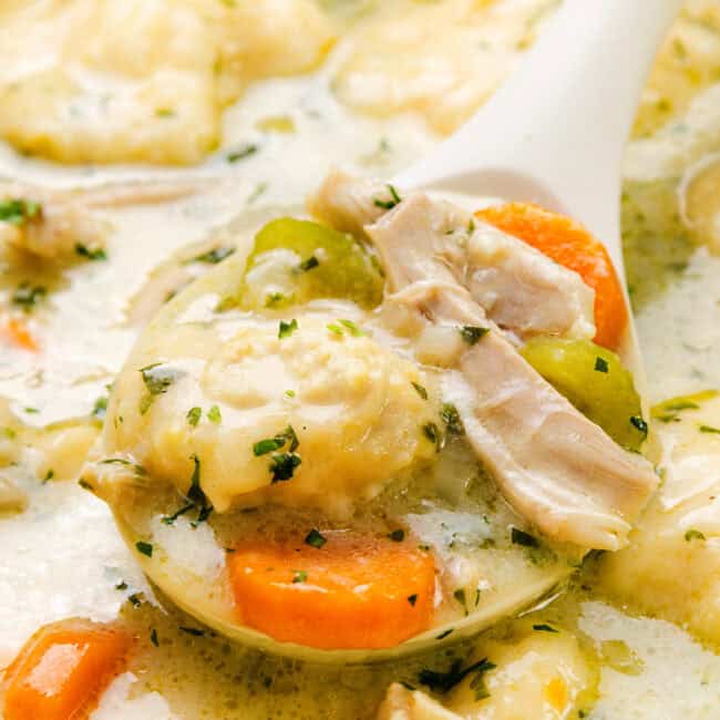 A close-up of creamy chicken and dumplings in a ladle reveals a delightful soup. The dish features tender chunks of chicken, carrots, and fluffy dumplings, garnished with herbs in a thick, savory broth. The background hints at more of the sumptuous stew waiting to be savored.