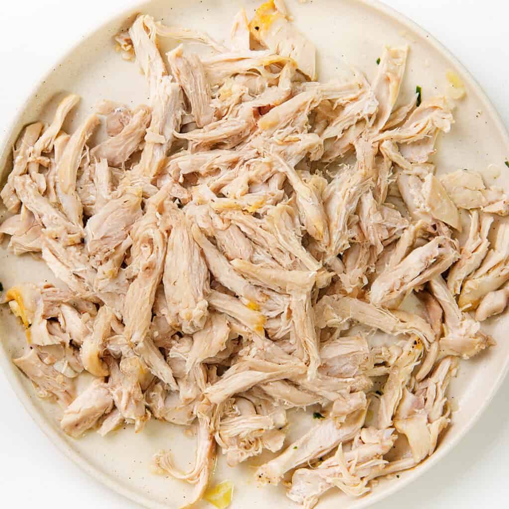 Shredded cooked chicken, perfect for a hearty Chicken Dumpling Soup, is piled on a round white plate. The chicken appears tender and juicy, with small bits of seasoning visible throughout.