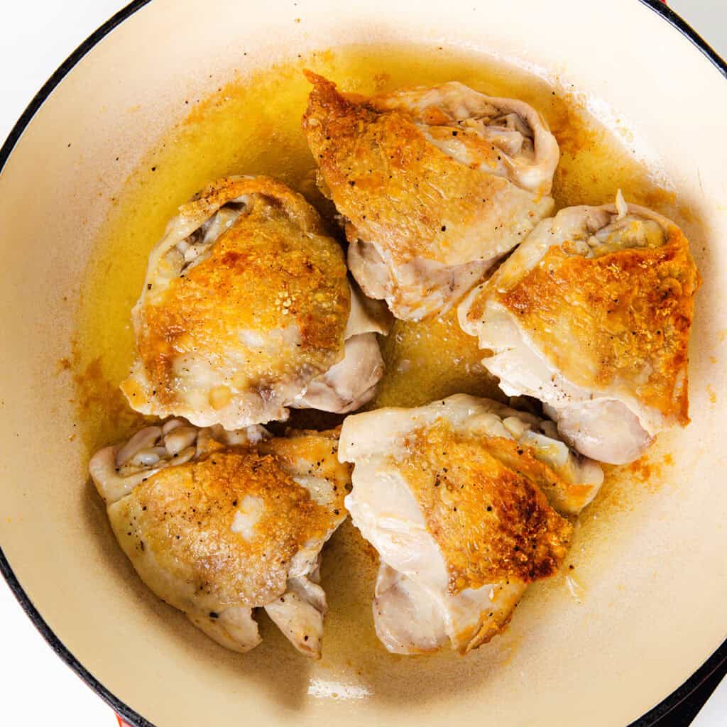 Five pieces of cooked chicken with crispy, golden-brown skin sit in a round pan with a hint of oil, reminiscent of the perfect base for a hearty chicken soup.