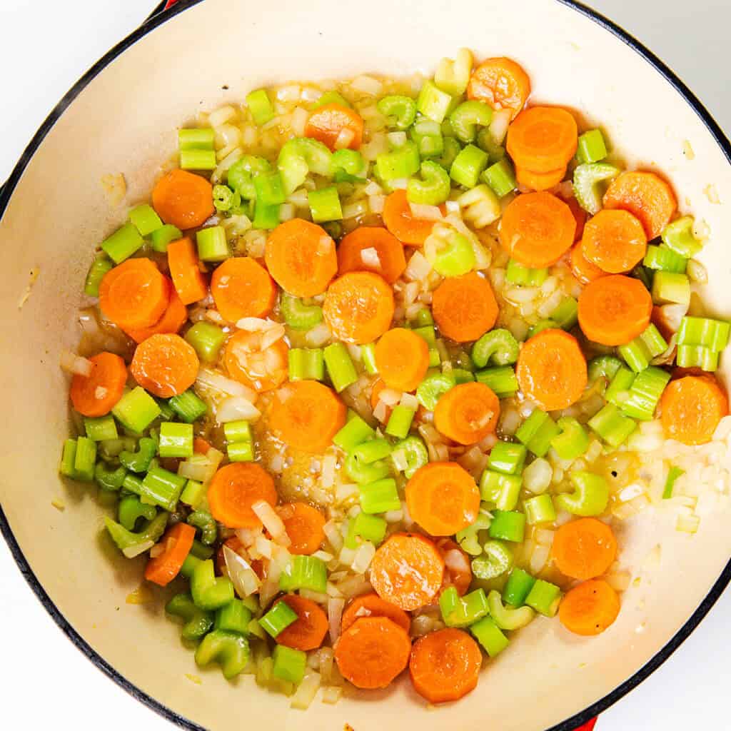 A pot filled with chopped carrots, celery, and onions is being sautéed. The vibrant vegetables mix seamlessly with tender chicken pieces, creating a colorful foundation for a hearty dumpling soup against the light background.