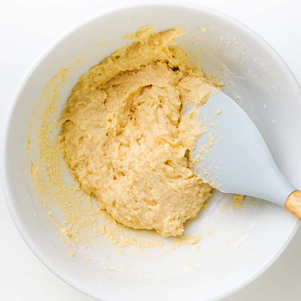 A white bowl filled with creamy batter, mixed with a light blue spatula resting on the edge, evokes the start of a delicious dumpling soup. The batter appears smooth with some lumps, ready for baking preparations.