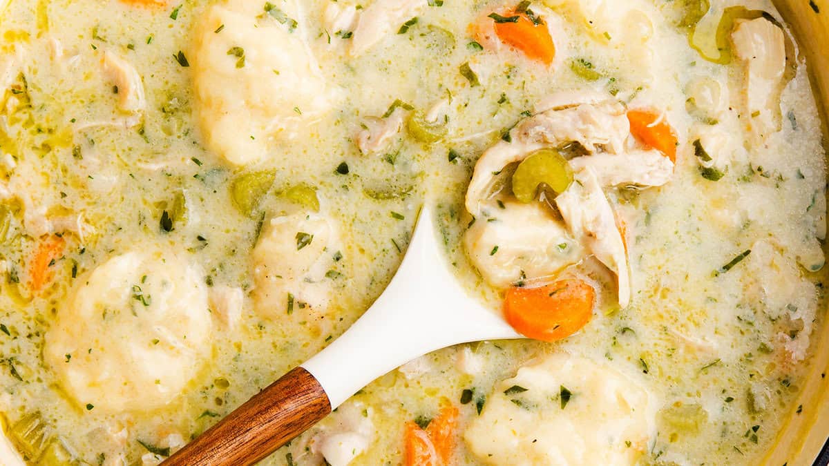 A creamy chicken and dumpling soup with chunks of chicken, carrots, celery, and herbs visible. A wooden-handled ladle scoops some of the thick soup, highlighting the tender dumplings and vibrant vegetables.