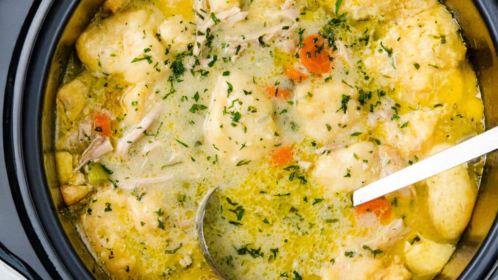 A close-up of Chicken Dumpling Soup in a pot showcases chunks of chicken, dumplings, sliced carrots, and chopped parsley in a creamy broth. A ladle and spoon are artfully placed inside the pot.