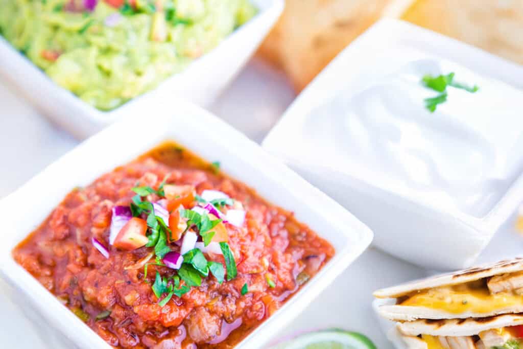 A close-up of three bowls containing guacamole, salsa with diced onions and cilantro, and sour cream. In the foreground, a slice of cheesy chicken quesadilla sits beside a lime wedge. The background shows tortilla chips.