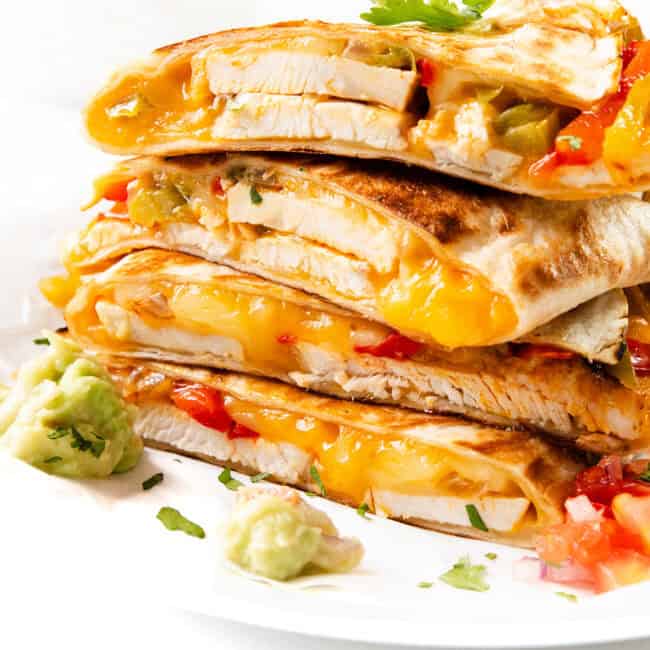 A stack of cheesy chicken quesadillas brimming with melted cheese, tender chicken, and vibrant vegetables sits on a white plate. Garnished with a sprinkle of cilantro, the dish is perfectly complemented by guacamole and diced tomatoes on the side.