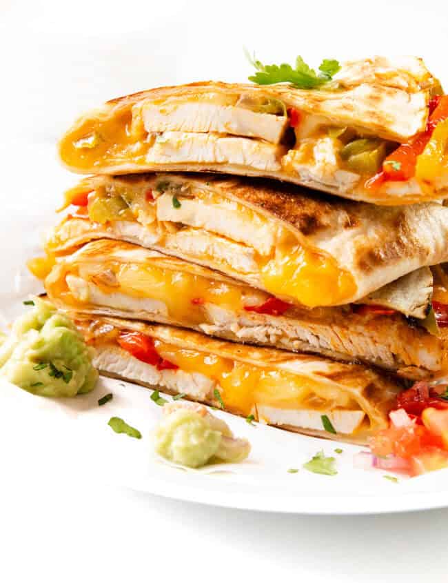 A stack of cheesy chicken quesadillas brimming with melted cheese, tender chicken, and vibrant vegetables sits on a white plate. Garnished with a sprinkle of cilantro, the dish is perfectly complemented by guacamole and diced tomatoes on the side.