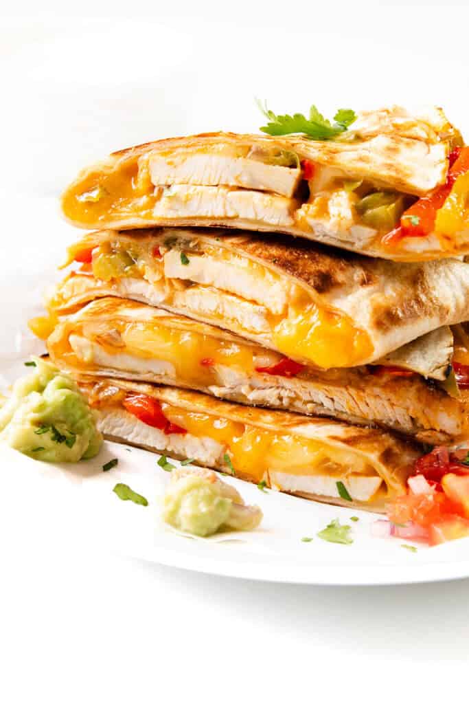 A stack of cheesy chicken quesadillas brimming with melted cheese, tender chicken, and vibrant vegetables sits on a white plate. Garnished with a sprinkle of cilantro, the dish is perfectly complemented by guacamole and diced tomatoes on the side.