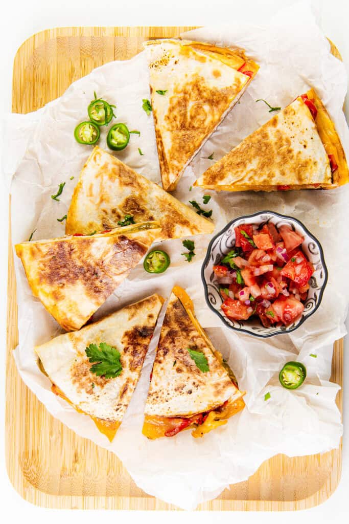 Sliced cheese quesadillas with grill marks are arranged on parchment paper. A small bowl of salsa accompanies them, garnished with sliced jalapeños and cilantro for a spicy kick.