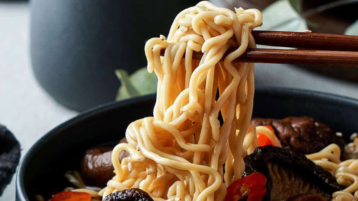 A bowl of noodles with mushrooms and red peppers, being lifted by chopsticks. The noodles appear cooked and are tangled around the chopsticks above the bowl. A dark, blurred background adds contrast to the focus on the food.