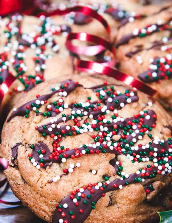 Festive Christmas cookies drizzled with chocolate and topped with red, white, and green sprinkles, surrounded by red ribbons and holly leaves with berries, adorn a wooden surface. Don't miss trying these delightful treats in 2024!