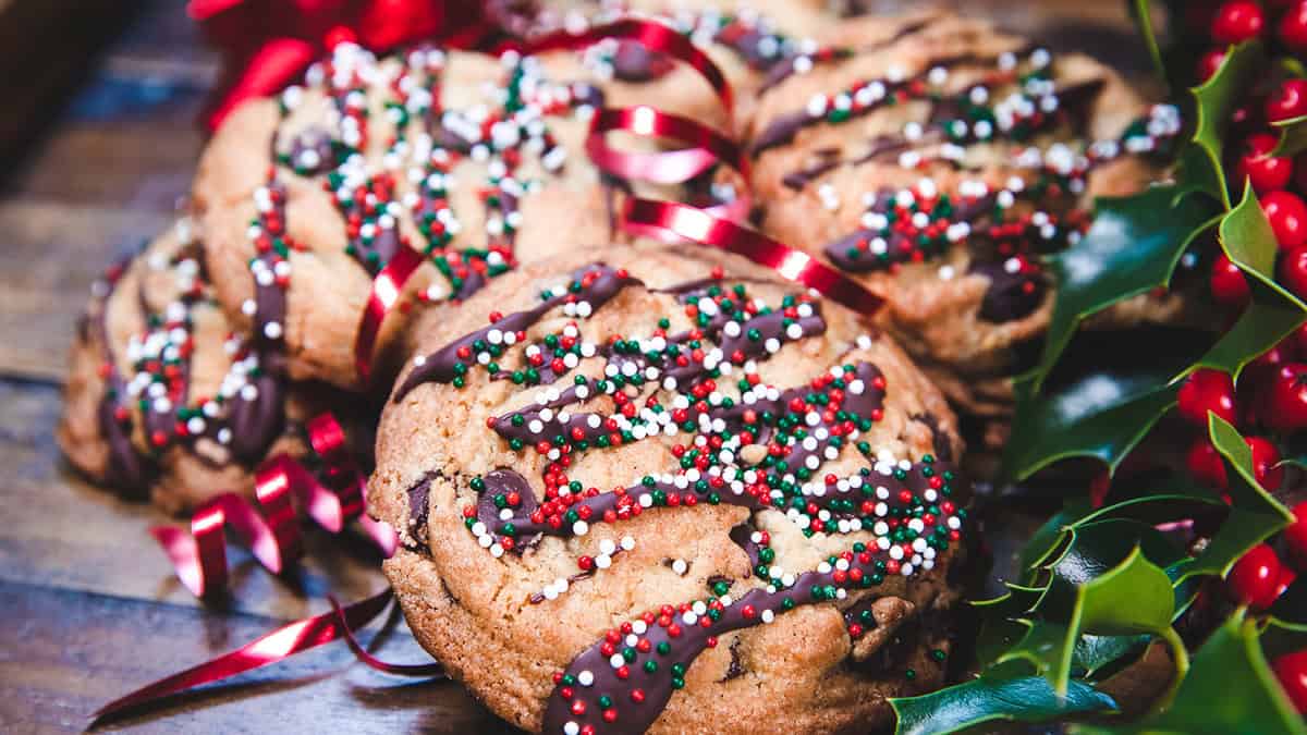 Festive Christmas cookies drizzled with chocolate and topped with red, white, and green sprinkles, surrounded by red ribbons and holly leaves with berries, adorn a wooden surface. Don't miss trying these delightful treats in 2024!