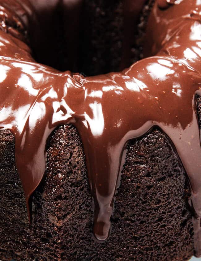 A chocolate bundt cake topped with glossy chocolate glaze, placed on a cooling rack. The rich glaze drips down the sides, highlighting the cake's moist and dense texture.