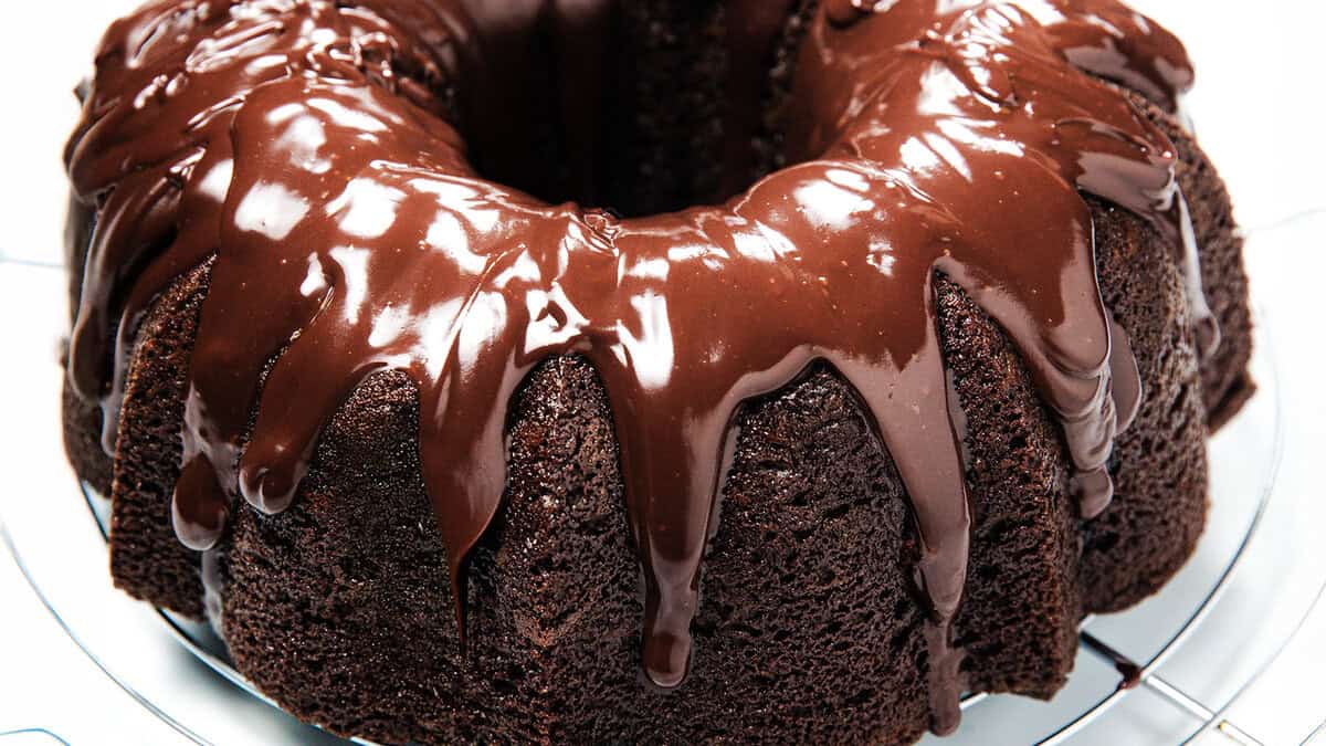 A chocolate bundt cake topped with glossy chocolate glaze, placed on a cooling rack. The rich glaze drips down the sides, highlighting the cake's moist and dense texture.