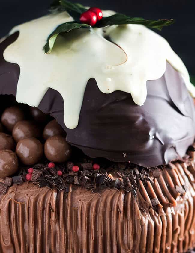 A festive cake with chocolate frosting and drizzled icing, topped with a large chocolate dome. The dome is partially broken, revealing chocolate balls inside. Reminiscent of Christmas cookies, it is garnished with holly leaves and berries, adding a touch of holiday spirit.