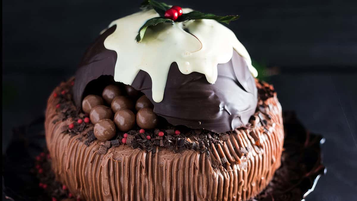 A festive cake with chocolate frosting and drizzled icing, topped with a large chocolate dome. The dome is partially broken, revealing chocolate balls inside. Reminiscent of Christmas cookies, it is garnished with holly leaves and berries, adding a touch of holiday spirit.