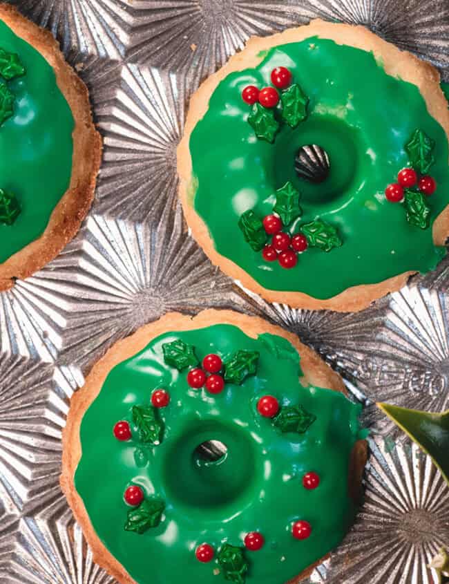 Six donut-shaped cookies with green icing and red candy decorations resembling holly berries are arranged on a textured silver surface.