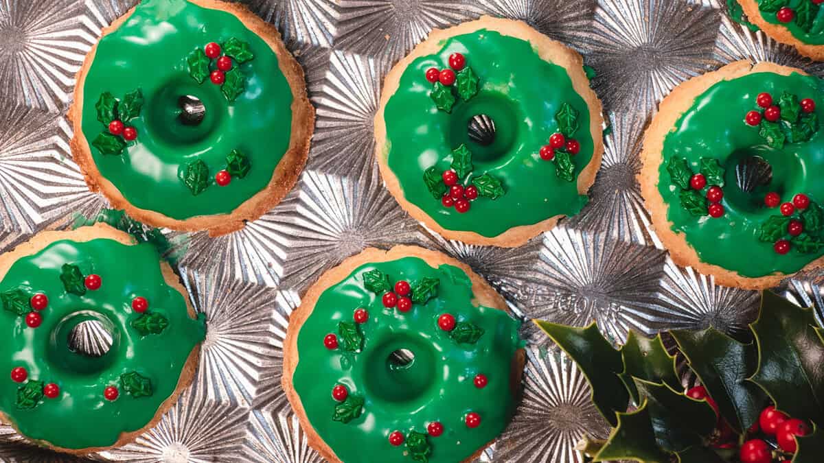 Six donut-shaped cookies with green icing and red candy decorations resembling holly berries are arranged on a textured silver surface.