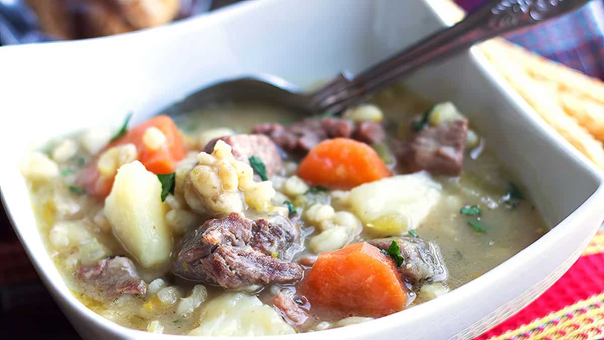 A close-up of a hearty, old school recipe bowl of soup features chunks of beef, carrots, potatoes, and barley in a rich broth. A spoon rests inside the square bowl. The background shows blurred elements, suggesting a cozy dining setting for enjoying classic dishes.