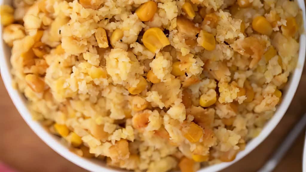 A close-up of a bowl filled with a mixture of bulgur wheat, corn kernels, and small cooked chickpeas. The dish appears grainy and textured, showcasing the different ingredients' yellow and light brown shades.