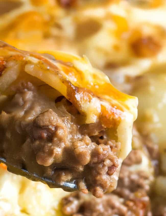 A close-up of a spoon serving a portion of creamy beef and potato casserole, showcasing layers of tender, browned ground beef and golden, melted cheese on top of thinly sliced potatoes in a baking dish.