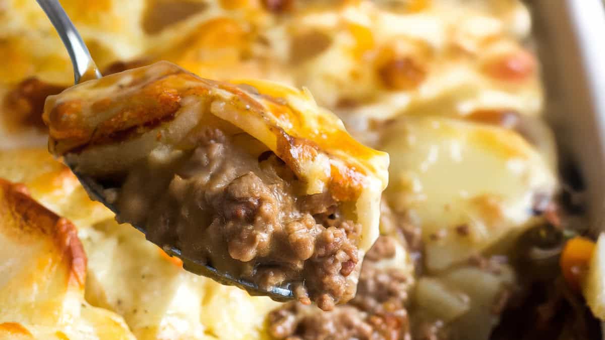 A close-up of a spoon serving a portion of creamy beef and potato casserole, showcasing layers of tender, browned ground beef and golden, melted cheese on top of thinly sliced potatoes in a baking dish.