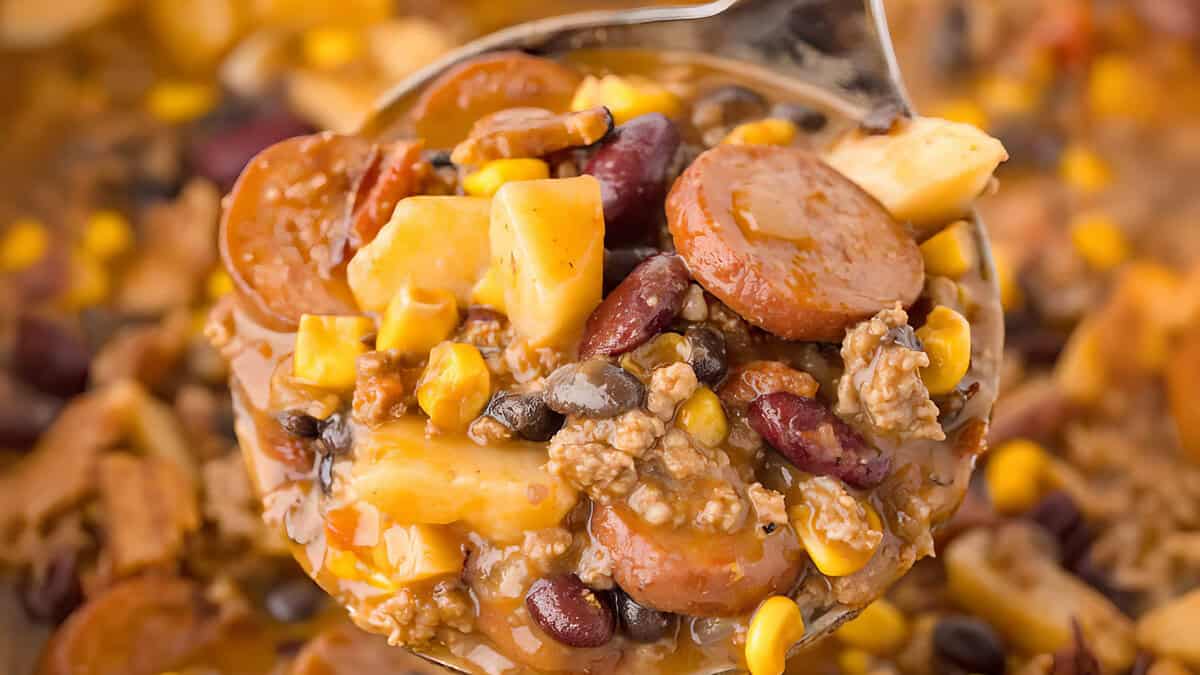 Close-up of a spoonful of hearty chili filled with sliced sausage, kidney beans, black beans, corn, cheese cubes, and ground beef in a rich, savory sauce.