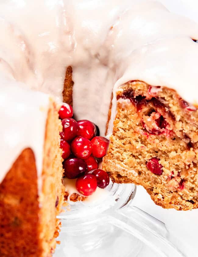 A sliced bundt cake with white icing on top, revealing a moist interior with red cranberries, showcases the allure of classic Bundt Cake Recipes. Presented on a clear glass stand, a cluster of bright red cranberries decorates the center for an inviting touch.