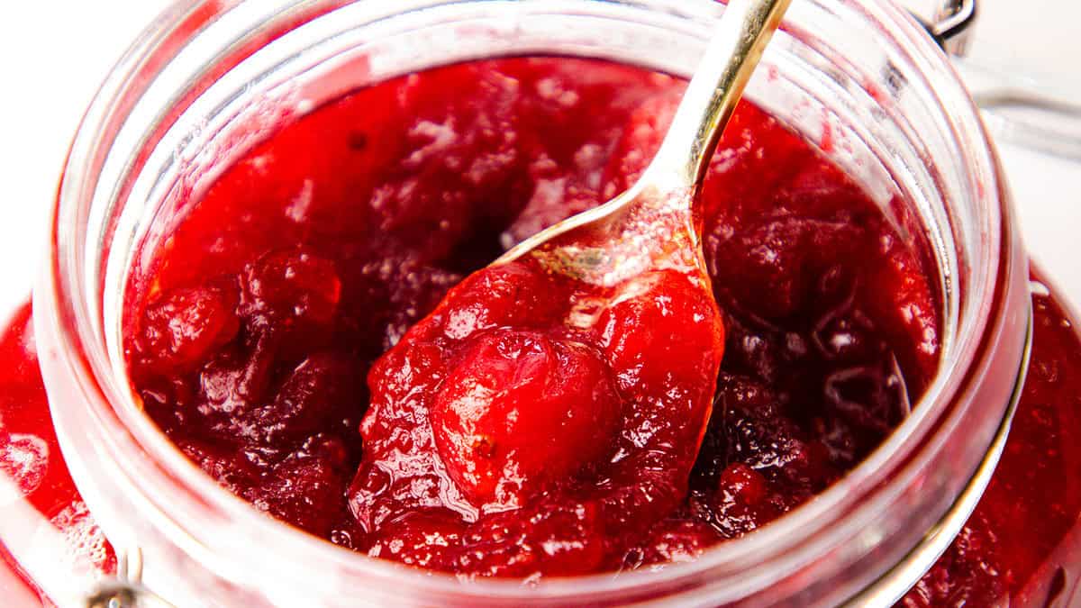 A close-up of a jar filled with vibrant red cranberry sauce. A spoon scoops into the textured, slightly chunky mixture, highlighting its glossy appearance. The jar is open, showing a metal latch on the side.