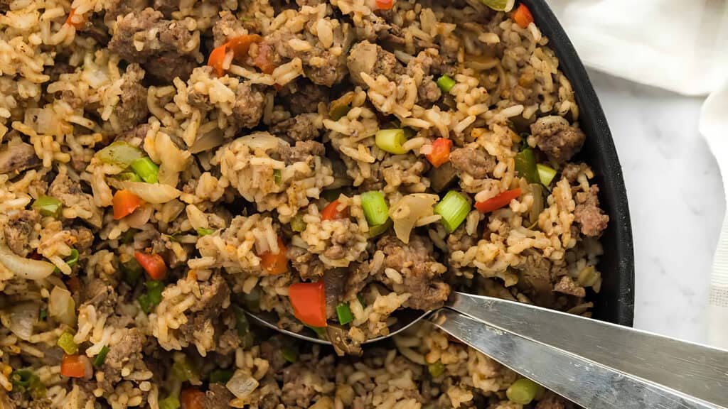 Close-up of a skillet filled with a savory rice dish, featuring ground meat, diced red peppers, green onions, and cooked rice. A shiny spoon is in the pan, ready to serve the meal.