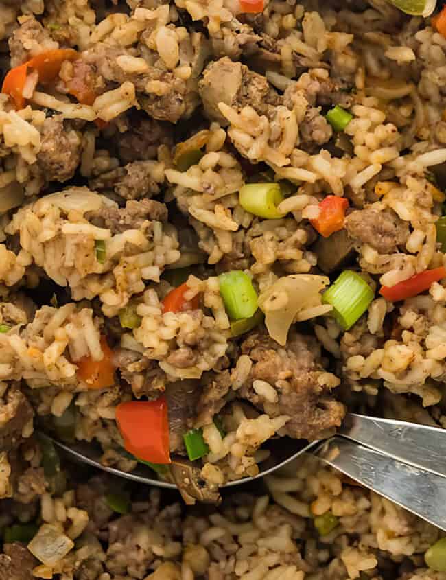 Close-up of a skillet filled with a savory rice dish, featuring ground meat, diced red peppers, green onions, and cooked rice. A shiny spoon is in the pan, ready to serve the meal.