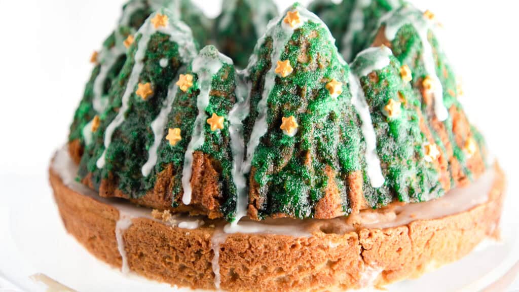 A festive cake designed to resemble a forest landscape, complete with tall, tree-like structures adorned with green icing and small star-shaped sprinkles. Drizzled with white icing to mimic snow, it pairs perfectly with Christmas cookies for a delightful holiday treat.