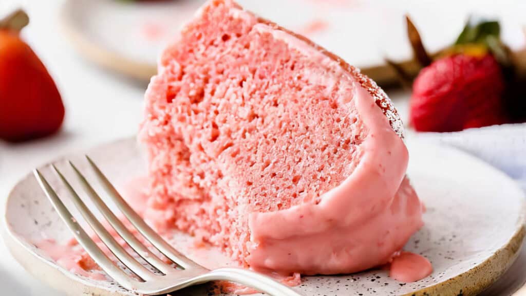 A close-up of a slice of pink strawberry bundt cake on a white plate, with creamy frosting and a fork beside it. In the background, blurred strawberries add a touch of red.