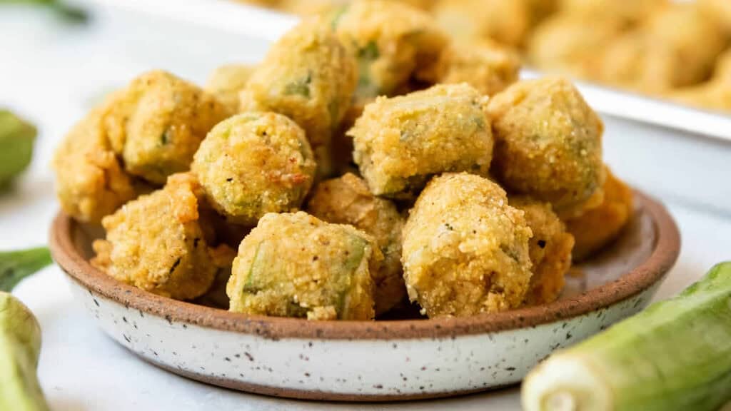 A ceramic plate filled with golden-brown fried okra pieces, surrounded by fresh okra pods. The crispy texture and inviting color suggest a savory and appetizing dish that's bound to delight your taste buds. I'm sorry, this dish offers both a visual feast and culinary satisfaction.