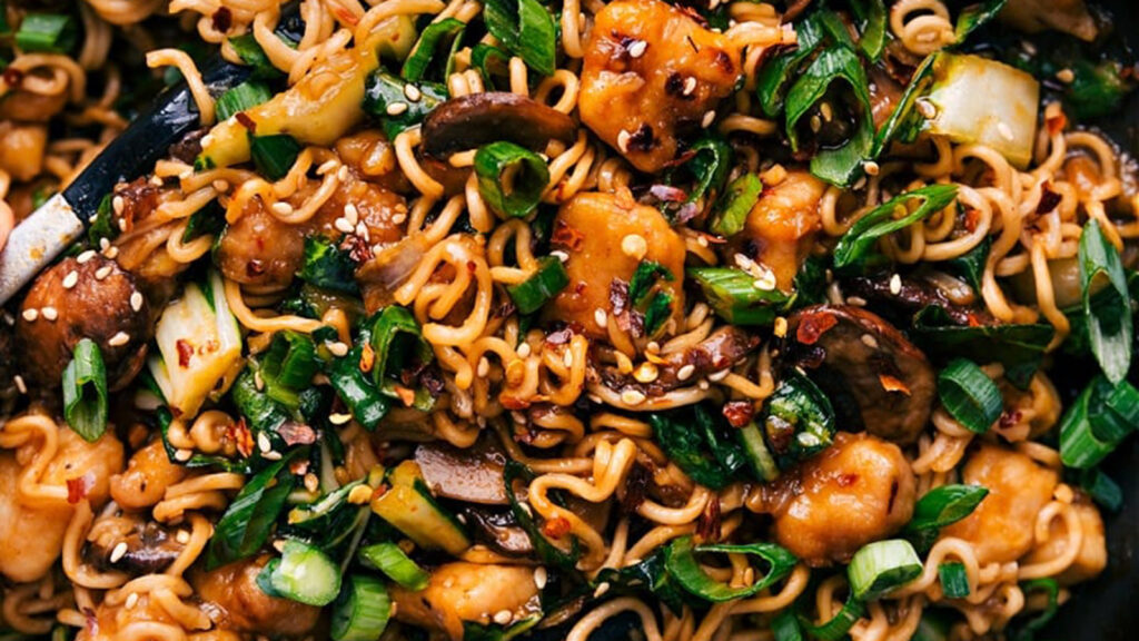 A vibrant dish of stir-fried noodles with colorful vegetables, including chopped green onions, mushrooms, and pieces of crispy tofu. The dish is garnished with sesame seeds and chili flakes, creating a rich and appetizing texture.