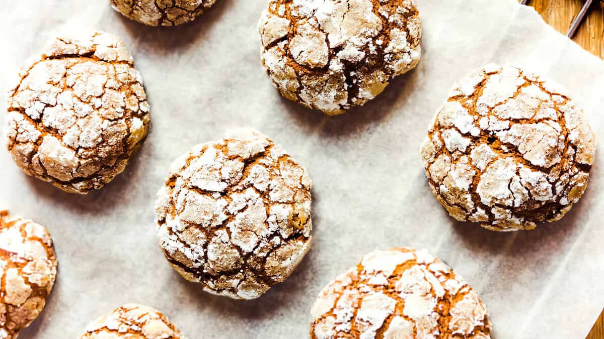 Crinkle cookies on a parchment-lined baking sheet. The cookies are round with a cracked surface, dusted with powdered sugar, giving a rustic and appealing appearance. They are arranged in rows, showcasing their golden brown texture.