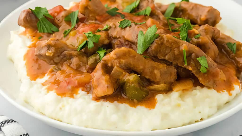 A plate of creamy grits potatoes topped with tender beef strips in a rich, savory gravy sauce, garnished with fresh parsley.