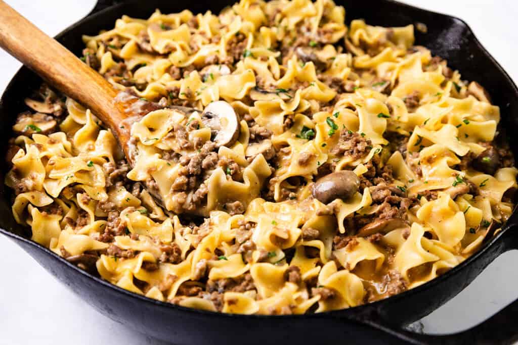 A skillet filled with one-pot beef stroganoff, featuring wide egg noodles, ground hamburger, sliced mushrooms, and a creamy sauce. Garnished with fresh herbs and stirred with a wooden spoon.