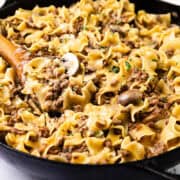 A skillet filled with one-pot beef stroganoff, featuring wide egg noodles, ground hamburger, sliced mushrooms, and a creamy sauce. Garnished with fresh herbs and stirred with a wooden spoon.