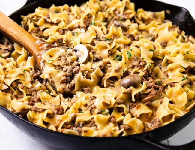 A skillet filled with one-pot beef stroganoff, featuring wide egg noodles, ground hamburger, sliced mushrooms, and a creamy sauce. Garnished with fresh herbs and stirred with a wooden spoon.