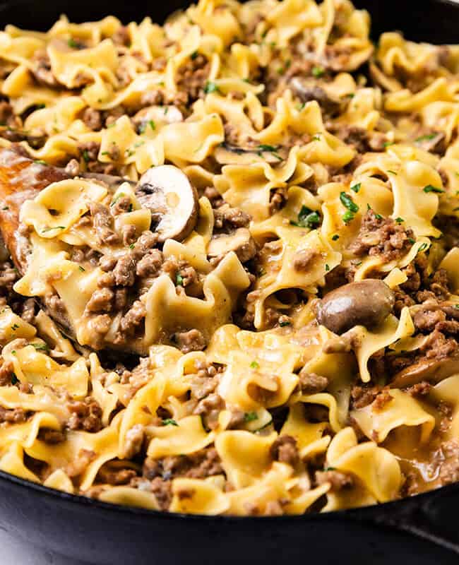 A skillet filled with one-pot beef stroganoff, featuring wide egg noodles, ground hamburger, sliced mushrooms, and a creamy sauce. Garnished with fresh herbs and stirred with a wooden spoon.