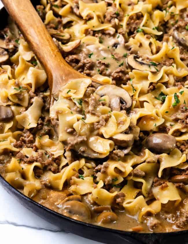 A skillet filled with creamy Hamburger Stroganoff, featuring broad egg noodles, slices of mushrooms, and ground beef, garnished with parsley. A wooden spoon is scooping a portion of the dish.