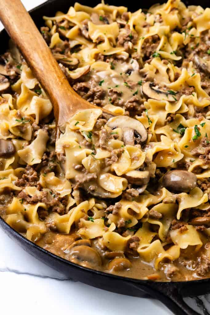 A skillet filled with creamy Hamburger Stroganoff, featuring broad egg noodles, slices of mushrooms, and ground beef, garnished with parsley. A wooden spoon is scooping a portion of the dish.