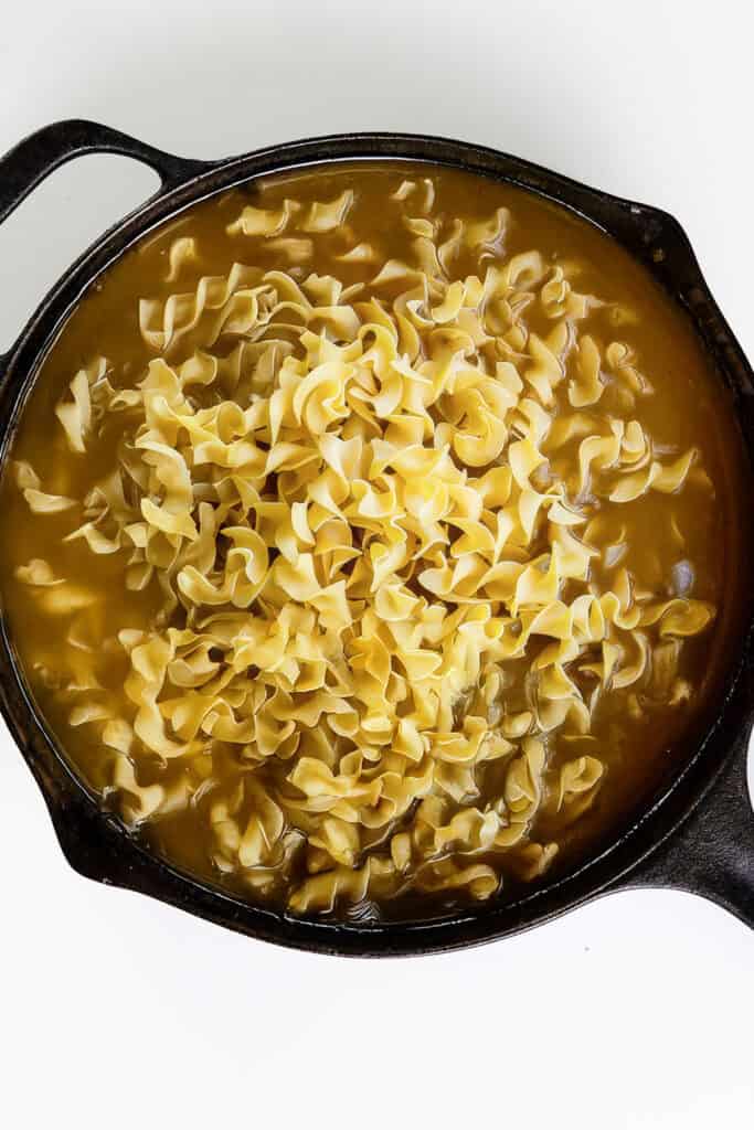 A cast iron skillet filled with uncooked egg noodles submerged in a light brown broth, ready to transform into a savory one-pot Stroganoff. The curly noodles are densely packed, covering most of the liquid's surface. The skillet rests on a pristine white background.