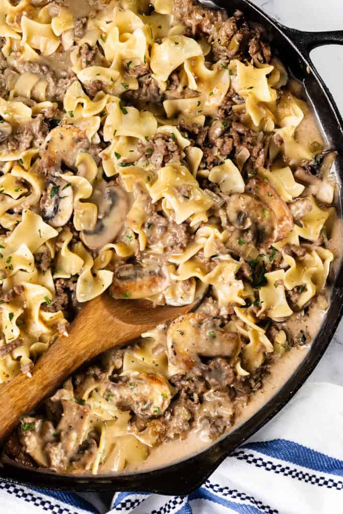 A skillet filled with creamy pasta featuring wide noodles, minced meat, mushrooms, and herbs in a savory sauce. This One Pot Hamburger Stroganoff is stirred by a wooden spoon, while a blue and white striped cloth peeks out from beneath the skillet.