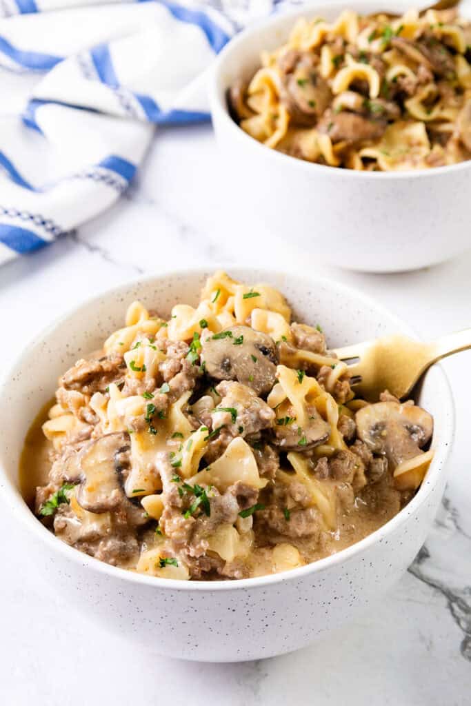 Two bowls of creamy hamburger stroganoff with egg noodles, mushrooms, and parsley garnish sit on a marble countertop. A gold spoon rests in the front bowl. A blue and white napkin is partially visible in the background, making this one-pot wonder both elegant and delicious.